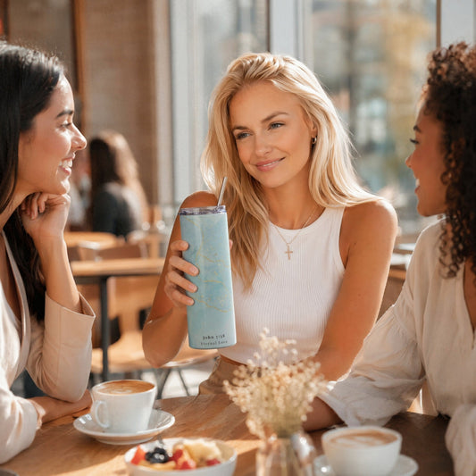 Woman holding a Living Waters stainless steel tumbler while talking with friends in a sunlit café, faith-inspired lifestyle scene.